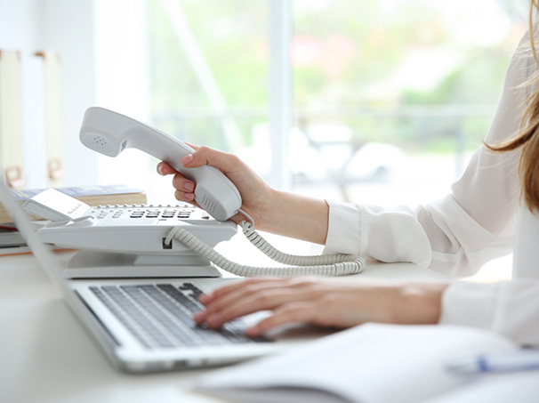 Person sitting at a desk picking up a phone with one hand and typing on a laptop with the other
