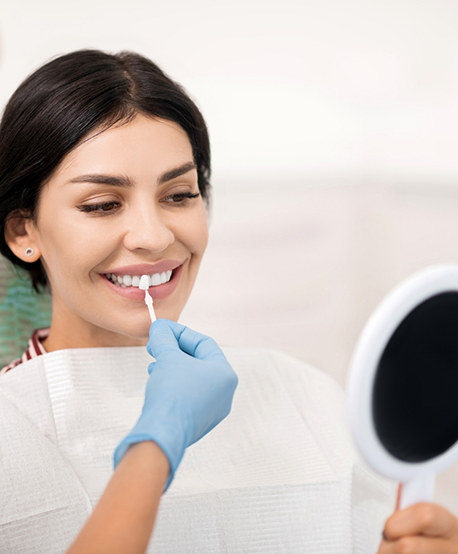 Dental patient looking at her smile in a mirror as her dentist holds a veneer in front of her tooth